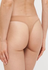 Beige thong ondergoed met een gladde textuur, minimalistisch ontwerp en dunne elastische tailleband, gezien vanaf de zijkant.