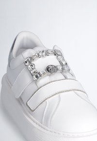 Sneaker bianco con tomaia in pelle liscia, doppie cinghie in Velcro, una fascia con accenti in argento e pietre preziose trasparenti, e suola in gomma testurizzata.
