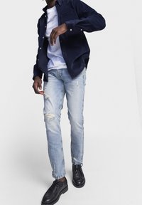 Marinblå kordflorsskjorta över vit t-shirt, kombinerat med ljusblå slitna jeans och svarta snörskor. Avslappnad outfit med lager på lager-texturer.