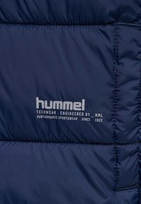 Marineblaue Puffermaterial mit glatter Textur, versehen mit weißen Druckschriftzügen. Das Material ist gesteppt mit sichtbaren Nähten und Logodetails.