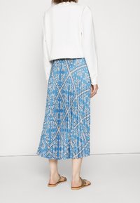 Nainen, jolla on päällään valkoinen pitkähiha, ja sininen punottu midi-hame, jossa on valkoinen paisley-kuvio ja ruskeat lapikkaat.