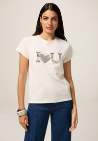 Camiseta de algodón blanca de manga corta, con un diseño de "Te ♥ U" en texturas negras y acentos plateados. Corte informal, combinada con vaqueros azul oscuro.