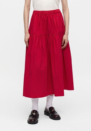 Personne portant une jupe midi rouge vif froncée, des chaussettes blanches, des mocassins noirs et un haut blanc, debout devant un fond blanc uni.