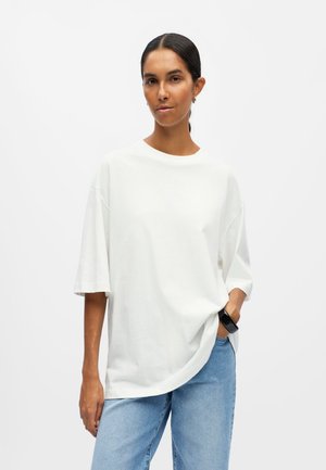 Femme aux cheveux foncés attachés portant un t-shirt blanc oversize, un jean bleu clair et une montre noire, debout devant un fond blanc uni.