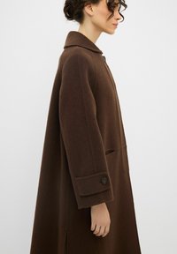 Cappotto di lana marrone con colletto abbottonato, tasche laterali e una vestibilità leggermente over-size. Presenta maniche arrotolate e un taglio dritto con una spacco all'orlo.
