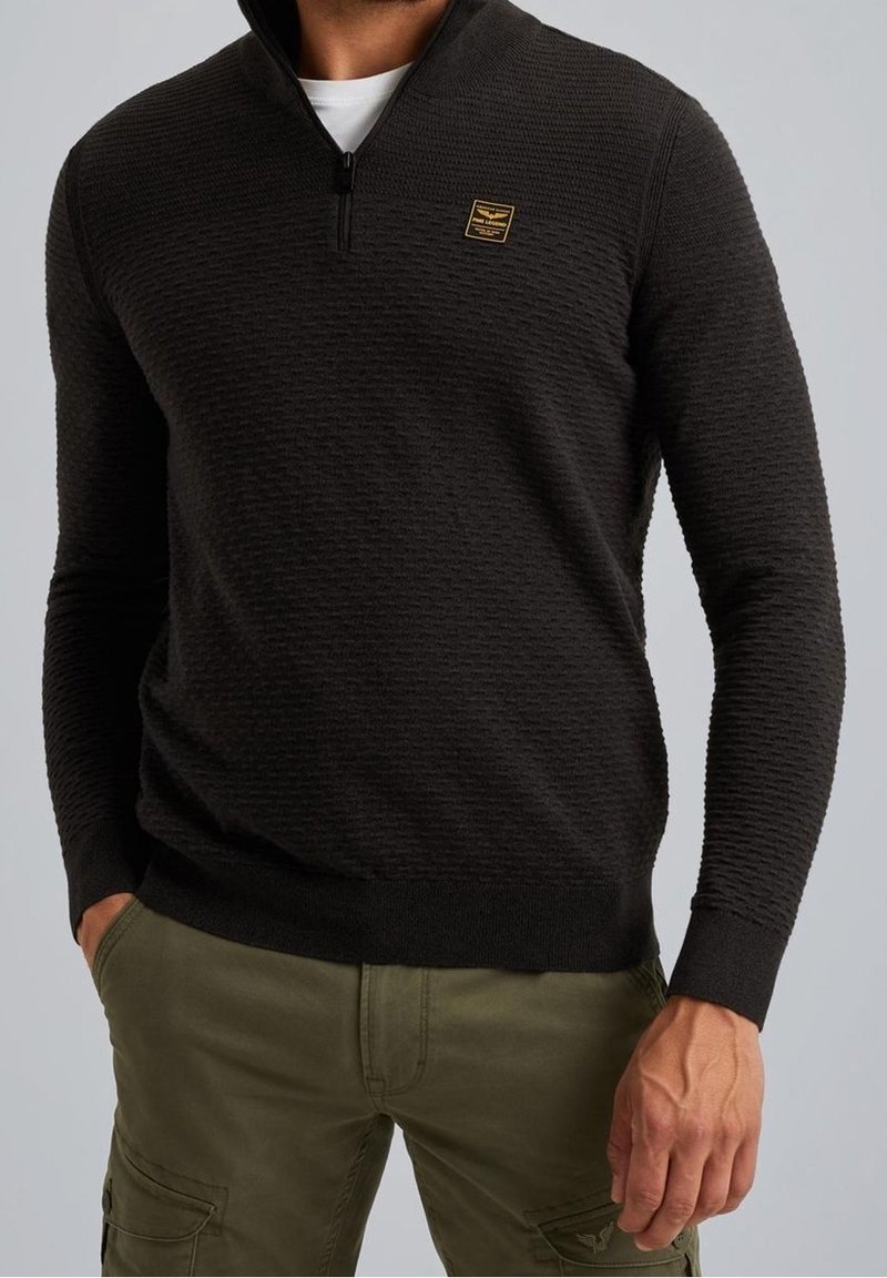Homme portant un pull noir texturé à demi-fermeture éclair et un pantalon cargo vert olive, les mains partiellement dans les poches, sur fond uni.