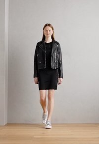 Svart skinnmotorsykkeljakke over en svart kjole, kombinert med metalliske sølv sneakers. Modellen går på tregulv mot en nøytral vegg.