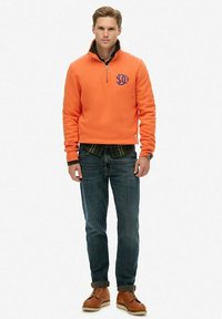 Orange quarter-zip pullover with embroidered logo, worn over a plaid shirt, paired with blue jeans and brown lace-up boots.