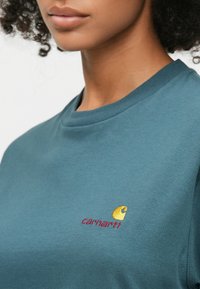 Τυρκουάζ βαμβακερό μπλουζάκι με στρογγυλή λαιμόκοψη. Διαθέτει κίτρινο κεντητό λογότυπο και κόκκινο κείμενο "carhartt" στην αριστερή περιοχή του στήθους. Λεπτή υφή.