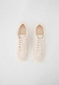 Lys beige sneakers med overdel i semsket skinn, rund tå, flate lisser og såle i hvitt gummi. Den teksturerte innersålen har små perforeringer.