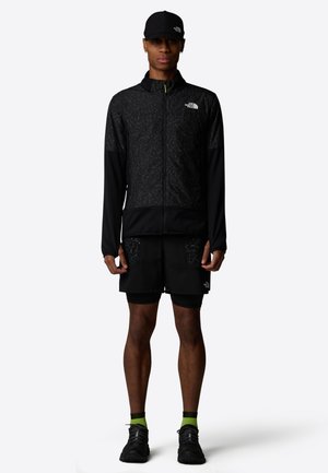 Chaqueta negra con cremallera y parte superior texturizada, combinada con pantalones cortos negros con bolsillos de parche. Zapatillas negras y calcetines con acentos verdes completan el look.