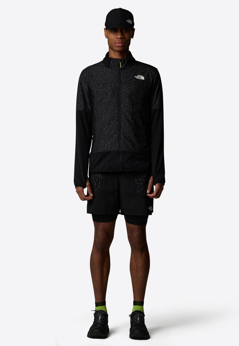 Chaqueta negra con cremallera y parte superior texturizada, combinada con pantalones cortos negros con bolsillos de parche. Zapatillas negras y calcetines con acentos verdes completan el look.
