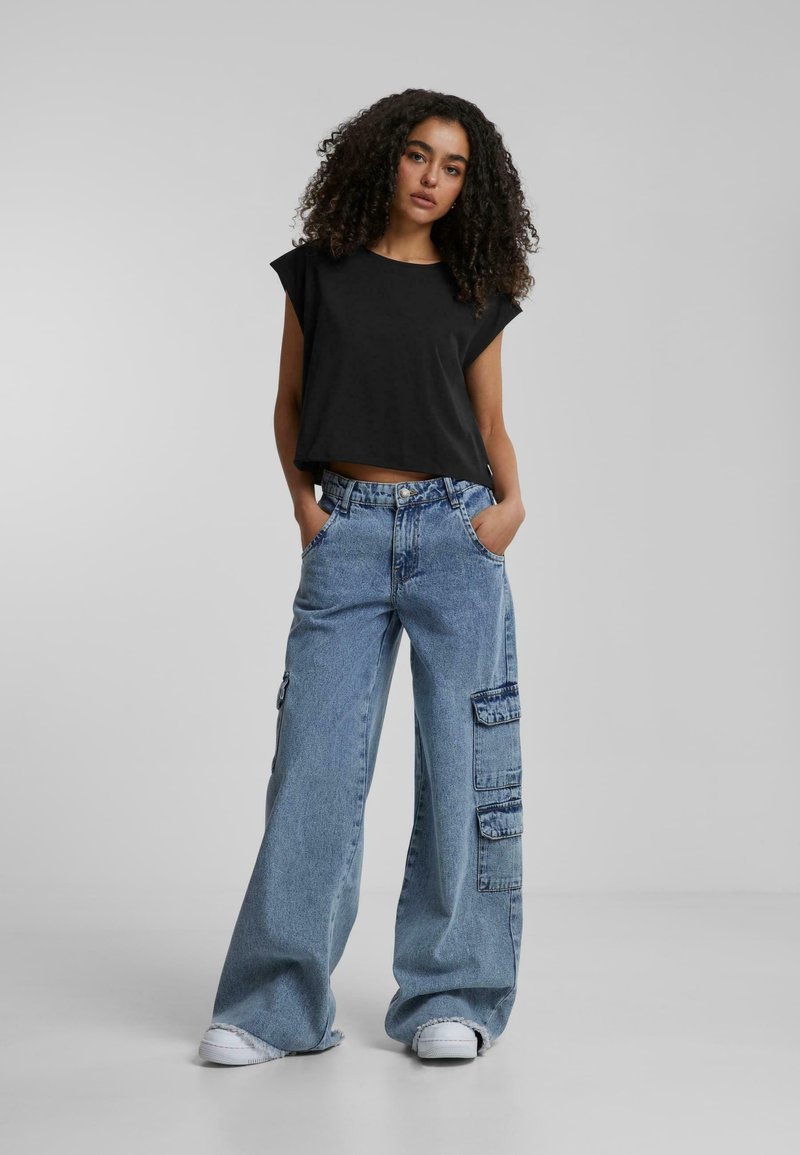 T-shirt noir court avec des manches larges, associé à un pantalon large en denim bleu clair comportant plusieurs poches et un ourlet effiloché. Baskets blanches.