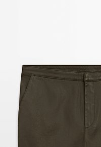 Section de pantalon vert olive foncé montrant la ceinture, la poche avant et la braguette devant avec des coutures visibles sur fond blanc.