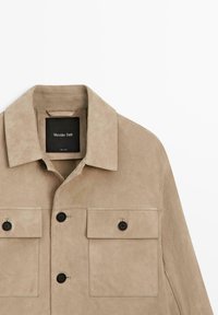 Beige Velourslederjacke mit weitem Kragen, zwei mit Knöpfen verschließbaren Brusttaschen, schwarzen Knöpfen und einem sichtbaren "Massimo Dutti"-Label innen am Kragen.