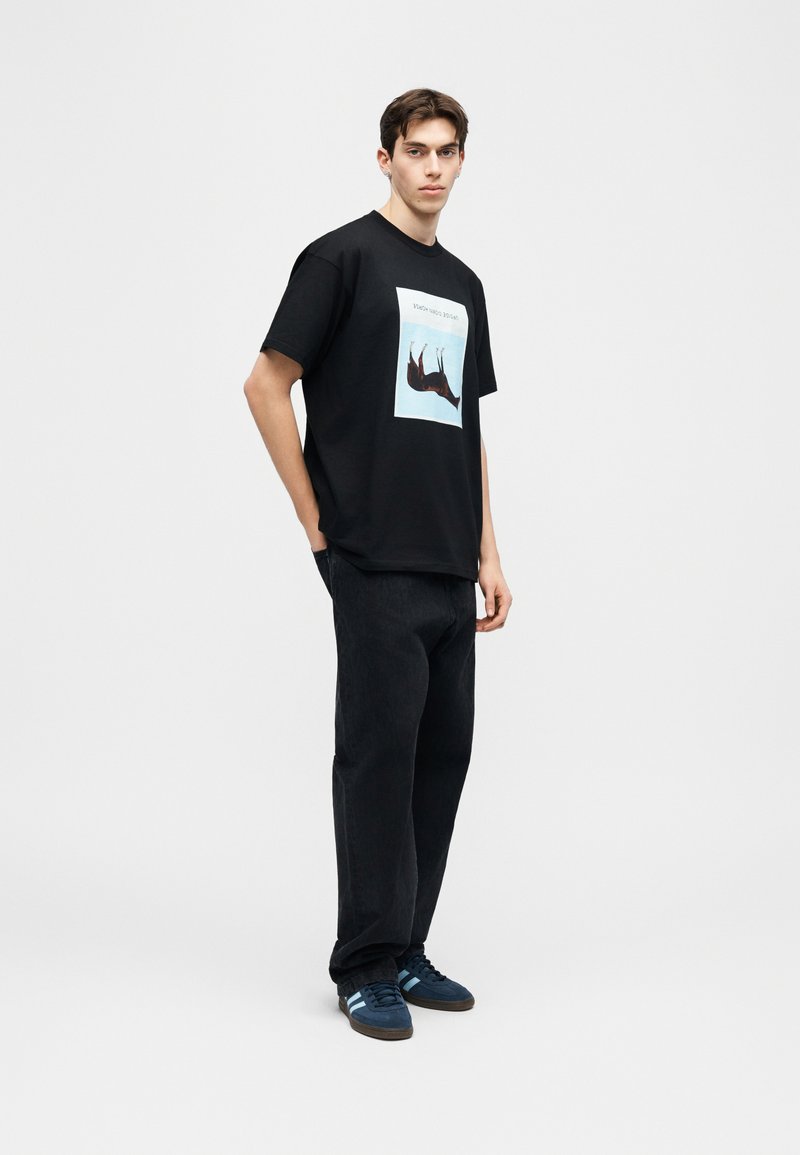 Junger Mann, der ein schwarzes T-Shirt mit Hirschmotiv, schwarze Hose und blaue Sneaker trägt, vor einem schlichten weißen Hintergrund steht.