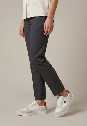 Person iført sorte bukser og hvide sneakers med røde hjerte detaljer, stående mod en neutral baggrund.