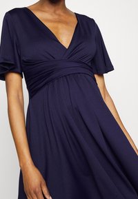 Mörkblå klänning med V-hals, korta fladdrande ärmar och en rynkad midja, tillverkad av ett mjukt, flödande tyg med en len textur.