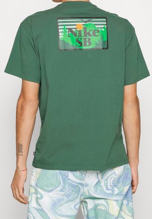 Osoba nosząca zieloną koszulkę Nike SB z logo inspirowanym naturą na plecach, w zestawieniu z niebiesko-zielonymi marmurkowymi szortami.
