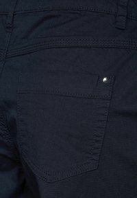 Pantalons en denim bleu marine avec une poche arrière dotée d'un rivet argenté. Le matériau semble solide avec des détails de couture visibles.