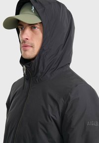 Schwarze isolierte Jacke mit hohem Kragen und Kapuze, ausgestattet mit einem Frontreißverschluss und Logodetails am Ärmel. Glattes, strukturiertes Material.