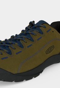 Chaussure de randonnée vert olive et bleu marine avec lacets bleus, semelle noire et logo visible de la marque Keen sur le côté.