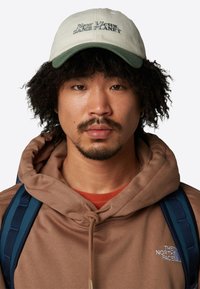 Gorra de béisbol con una corona verde claro y un visor verde más oscuro, que presenta el texto "Nuevas Perspectivas. El mismo PLANETA." Sudadera marrón y correas de mochila visibles.