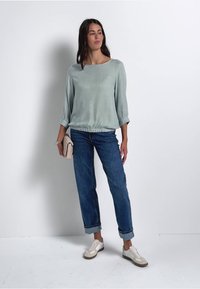 Blusa verde chiaro con una vestibilità rilassata e orlo elasticizzato, abbinata a jeans blu medio e sneakers bianche con dettagli beige.