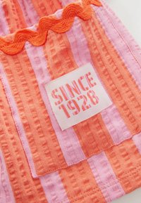 Tissu texturé rayé orange et rose avec un écusson cousu portant la mention "Since 1928" et une bordure ondulée orange en haut.