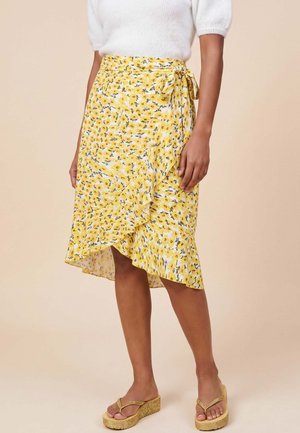 Vrouw die een gele bloemenwikkelrok met ruches draagt, een witte trui met korte mouwen en gouden platformslippers tegen een beige achtergrond.