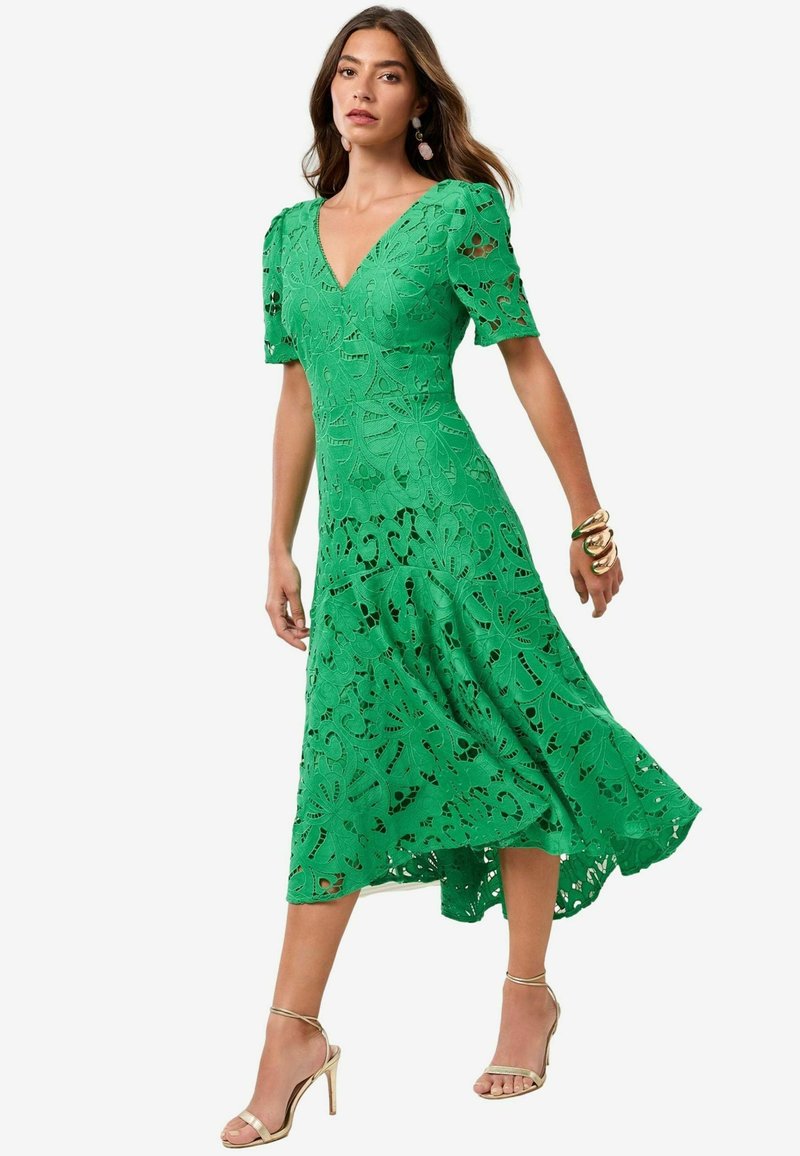 Rochie verde din dantelă cu decolteu în V, mâneci scurte, modele florale decupate și un tiv flared, asimetric. Asortată cu sandale aurii cu bretele.