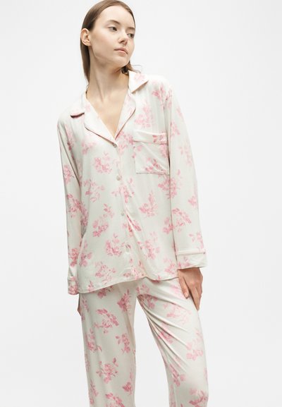 Femme portant un pyjama blanc à manches longues boutonné avec des motifs floraux roses, debout devant un fond blanc uni.
