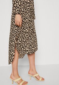 Robe midi à imprimé léopard beige avec accents noirs, dotée d'une fente latérale. Associée à des sandales à talons hauts jaunes clair à double bride.