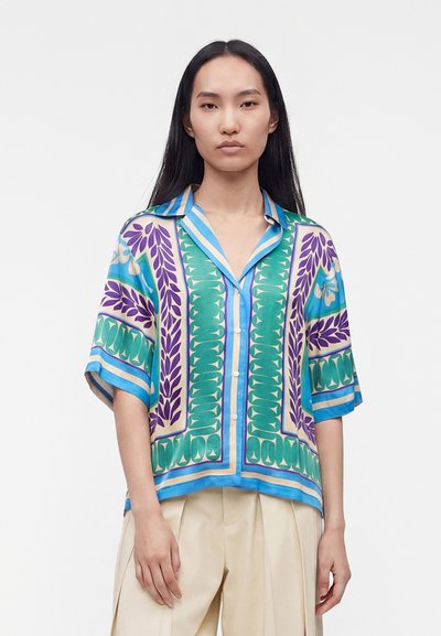 Femme aux longs cheveux noirs porte une chemise ample à manches courtes avec des motifs floraux bleus, verts et violets et un pantalon beige à jambes larges.