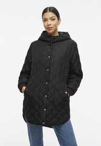 Črn prešiten plašč s kapuco, zaponko na gumb, stranskimi žepi in rebrastimi manšetami. Ima dolg, sproščen kroj. Gladek videz.