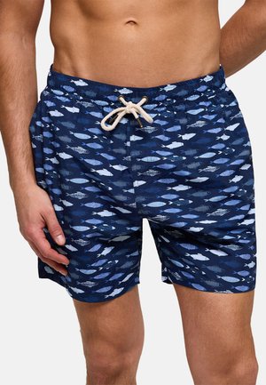 Hombre vistiendo pantalones cortos de baño azul marino con patrones de peces en azul claro y blanco, y un cordón blanco, de pie con una mano relajada.