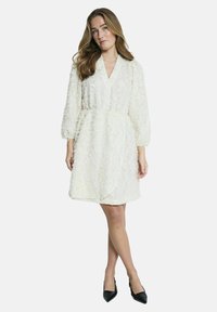 Witte textuur jurk met een V-hals, lange pofmouwen, aangesnoerde taille en een asymmetrische zoom. Gepaard met zwarte hoge hakken.