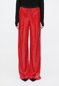 Personne portant un pantalon à paillettes rouge vif, large, et un haut noir à manches longues, debout sur un fond blanc uni.