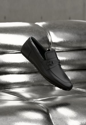 Mocassin en cuir noir avec détail boucle reposant sur une surface argentée métallique et capitonnée réfléchissante.