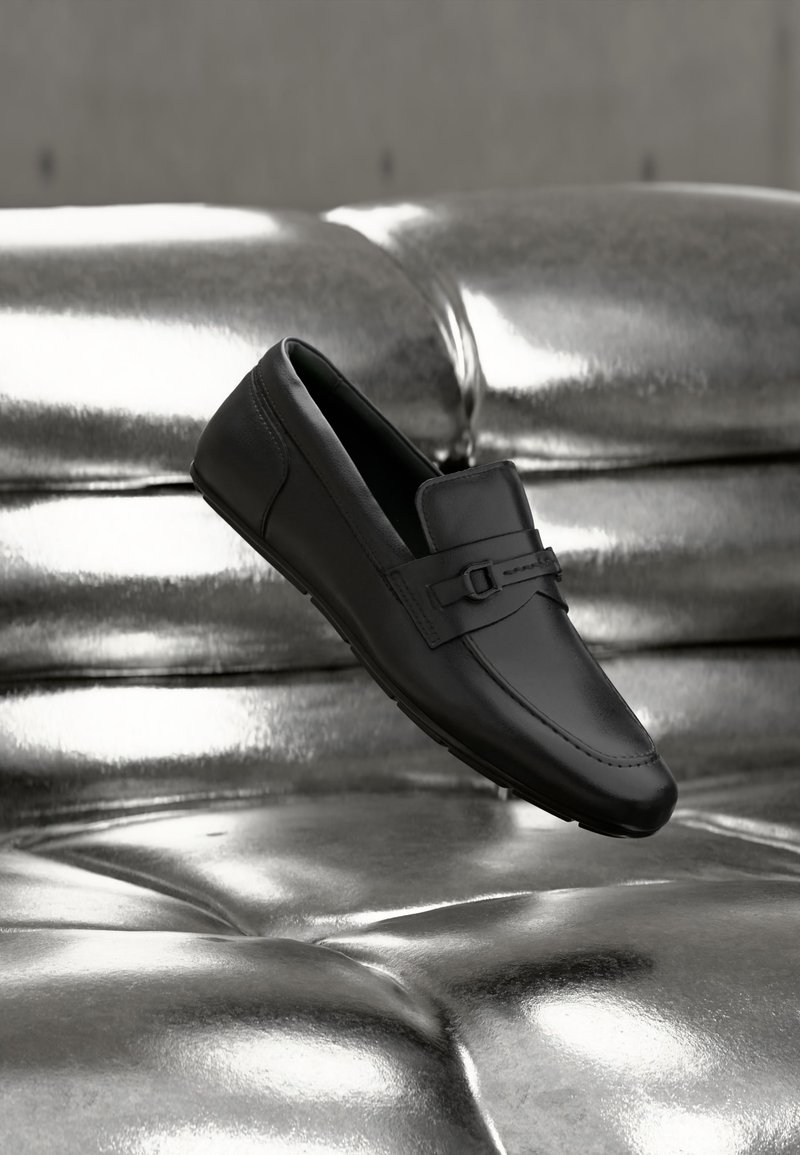 Mocassin en cuir noir avec détail boucle reposant sur une surface argentée métallique et capitonnée réfléchissante.