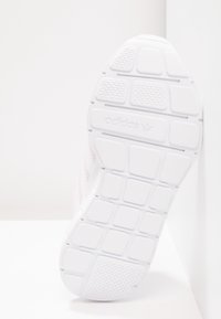 Bílá podrážka tenisky Adidas s texturovanými gripovými vzory a logem Adidas umístěným uprostřed podrážky, pořízená na bílém pozadí.