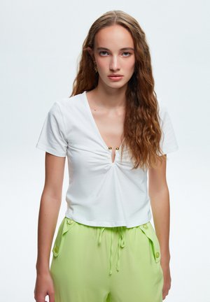 Hvid cropped T-shirt med V-udskæring og samlingdetaljer med to gyldne accenter. Modellen bærer limegrønne bukser med snørebånd og knapper.