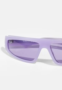 Lila műanyag napszemüveg, letisztult dizájnnal, körbeölelő formával és sötét árnyalatú lencsékkel; Ray-Ban logó látható a kereten.