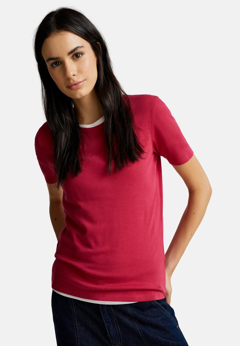 Giovane donna con lunghi capelli scuri che indossa una maglietta rossa a maniche corte sopra una canotta bianca e pantaloni di denim scuri, in posa su uno sfondo bianco.