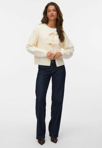 Krämfärgad k kardigan med en kortad design, ribbade manschetter och två knytband. I kombination med mörkblå vida jeans och spetsiga bruna skor.