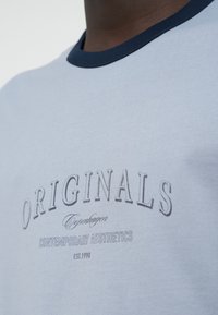 Ανοιχτό μπλε T-shirt με αντίθετη σκουρόχρωμη μπλε ριμπ λαιμόκοψη, με κείμενο «ORIGINALS Copenhagen» σε σκούρο γκρι και «CONTEMPORARY AESTHETICS» από κάτω.