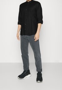 Camicia nera button-up con colletto cinese, abbinata a jeans grigi arrotolati e sneakers nere con suola spessa.