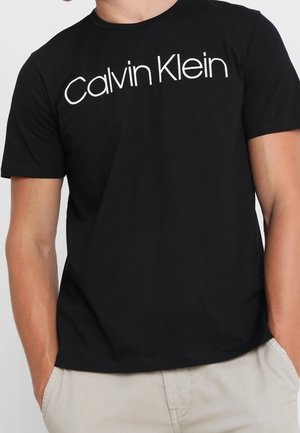 Sort kortærmet t-shirt med "Calvin Klein" skrevet i hvidt tværs over brystet, båret med lysebeige bukser og hænder i lommerne.