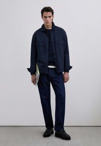 Navy shirt jacket with two chest pockets over a dark sweater, paired with dark jeans and black shoes. Model holding a magazine.