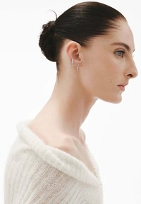Pendientes de plata y oro con diseños intrincados colgando de la oreja, complementados por un suave suéter blanco de punto sin mangas.